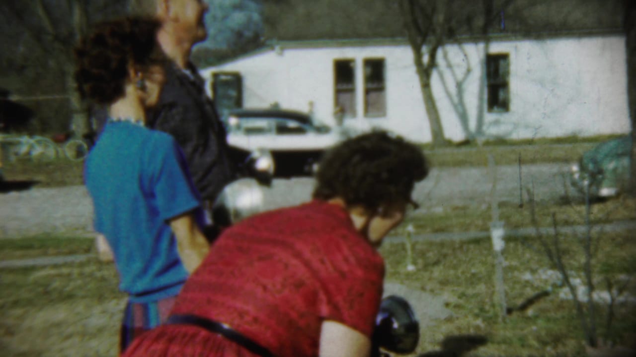 1959 Chadron, Nebraska home-movie frame showing a backyard group, a woman in red bending down, a girl in blue, and a white car near a house.