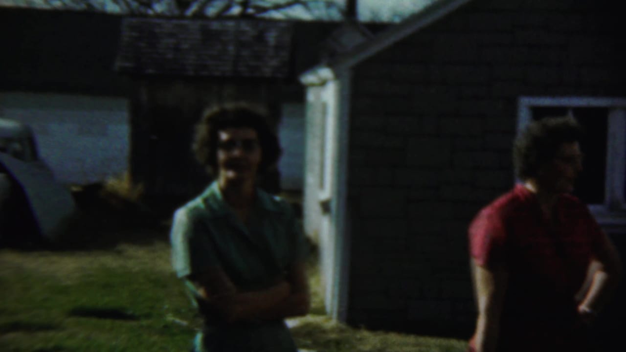 Two women stand near a dark house wall, with a white car and outbuilding in the background.
