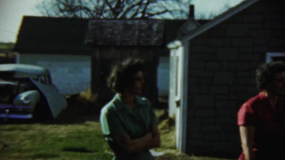 1959 Chadron, Nebraska backyard frame showing a woman in green beside house siding, with a white midcentury car partly visible behind her.