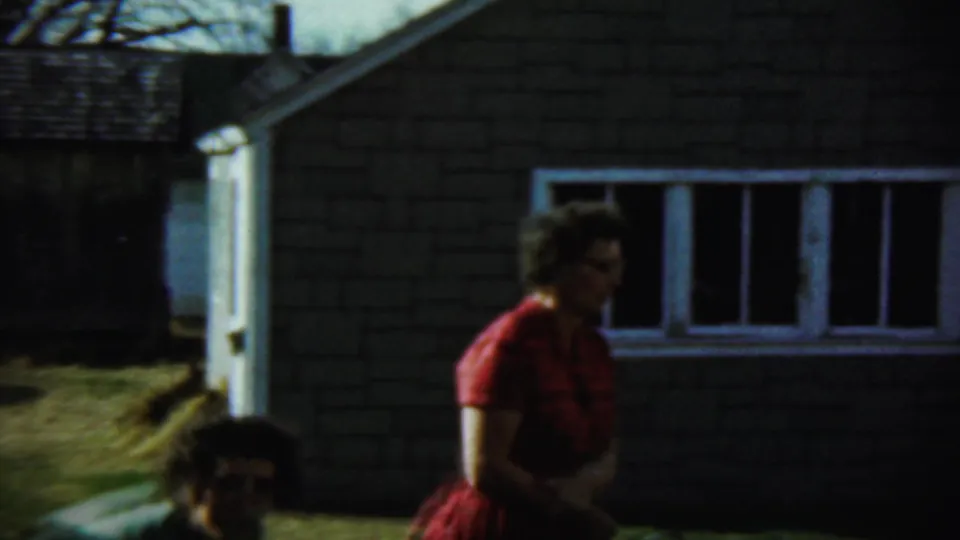 1959 Chadron, Nebraska frame showing a woman in red standing near a brick-sided house and white-framed window in the backyard.