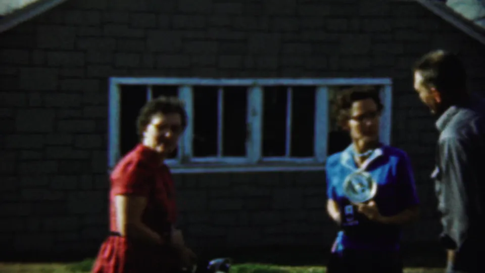 In Chadron, Nebraska, 1959, two women in colorful dresses hold flash cameras while a man stands nearby outside a windowed building.