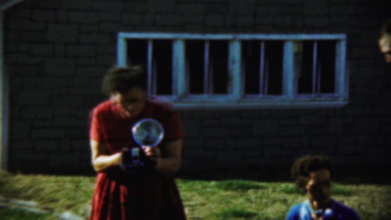 A woman in a red dress examines a flash camera near another seated figure.