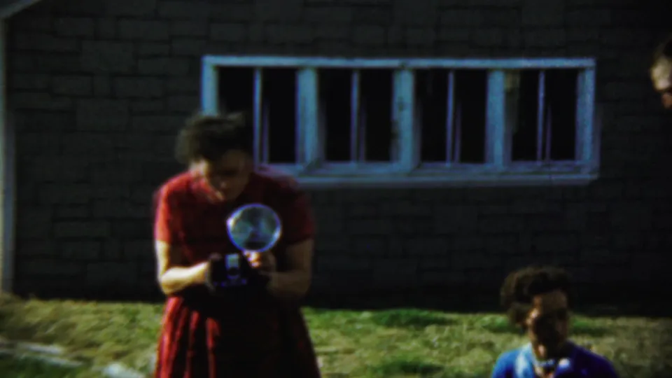 A woman in a red dress looks down at a vintage flash camera outside the Chadron building, with another person partly visible nearby.