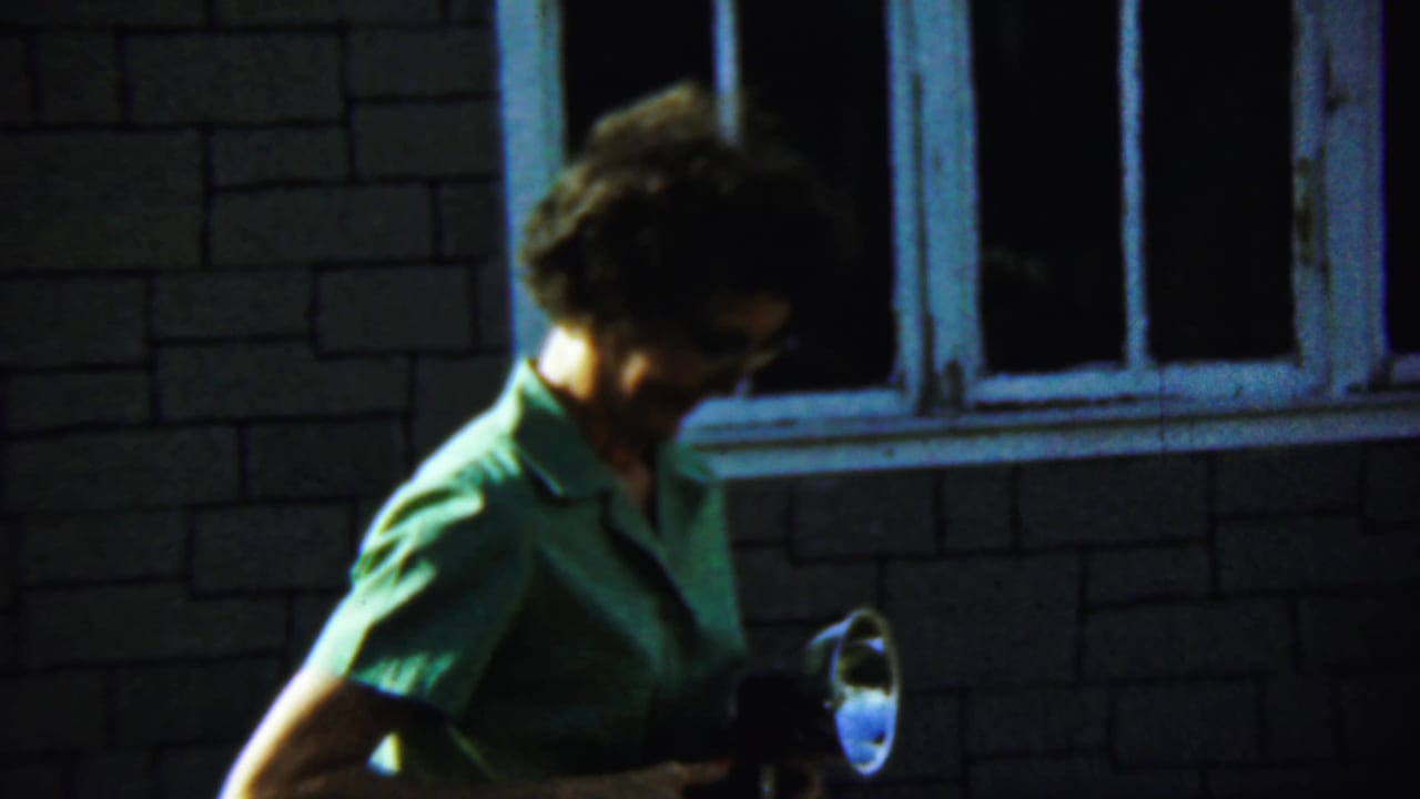 In Chadron, Nebraska, 1959, a woman in a green collared shirt looks down while holding a flashlight outside a brick building.