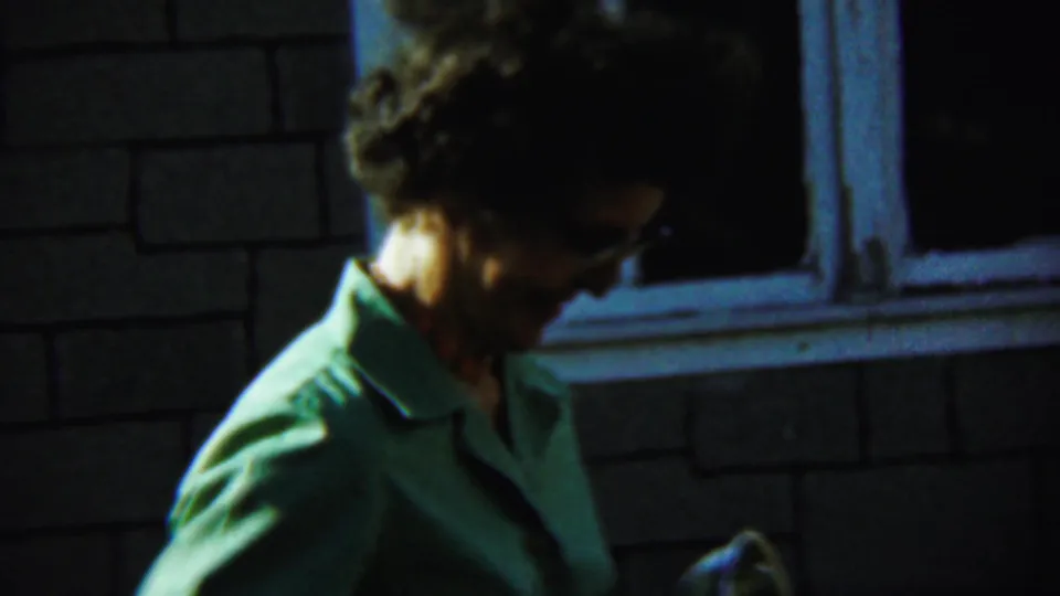 In the 1959 Chadron home-movie frame, the woman lowers her gaze near the brick wall and dark window opening.
