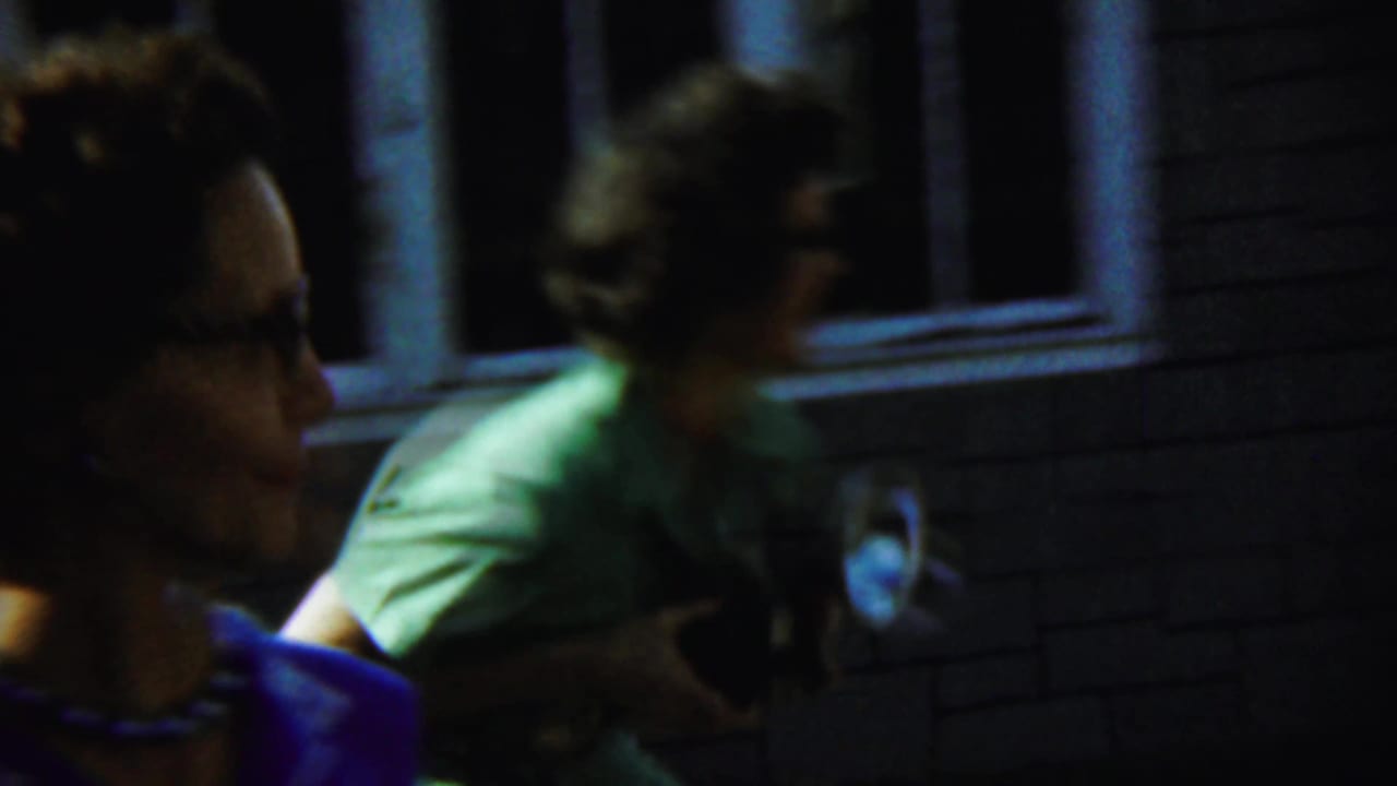 Two women move past a brick house, one close to camera and one carrying a camera.