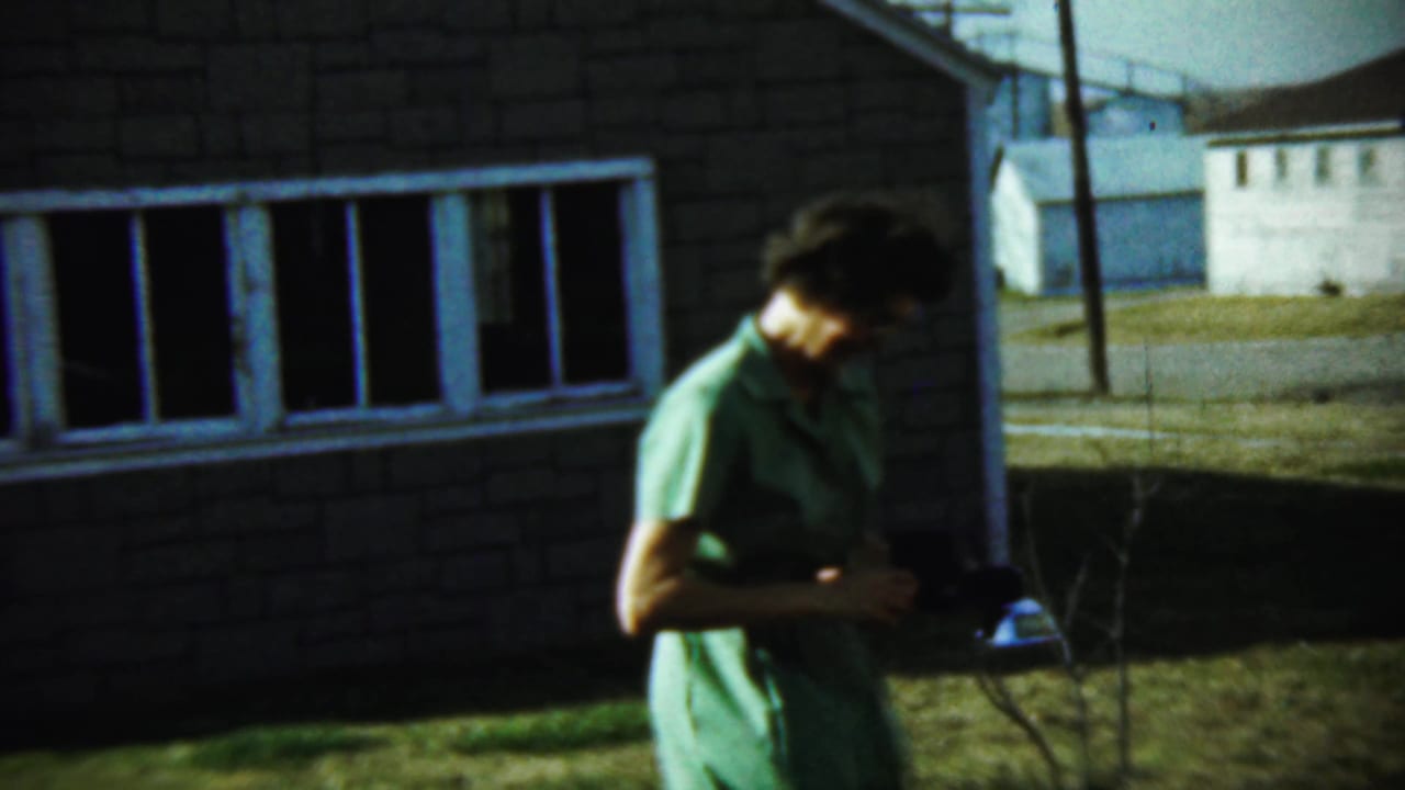 Woman in a green dress stands beside a brick house holding a camera.