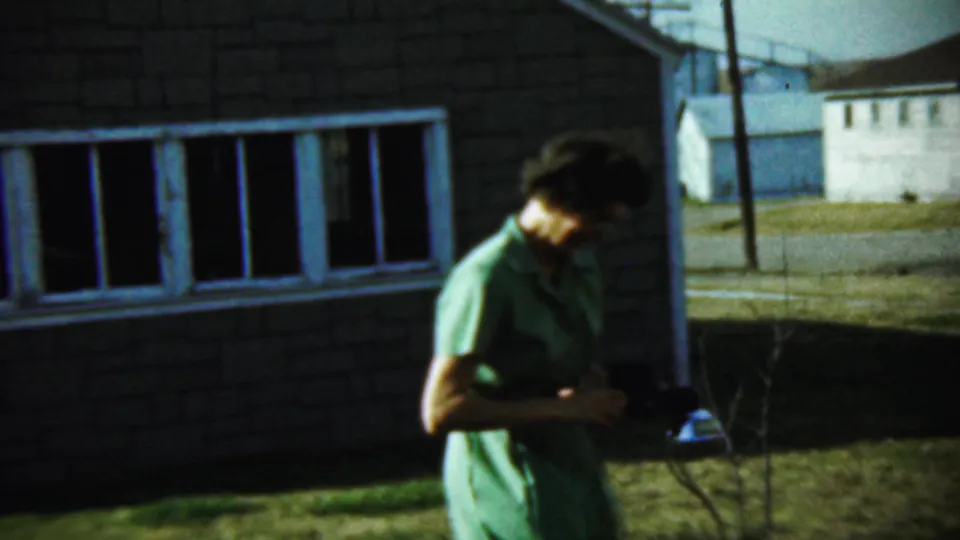 A woman in a green dress stands on a lawn in Chadron, Nebraska, holding a camera near a brick house in 1959 footage.