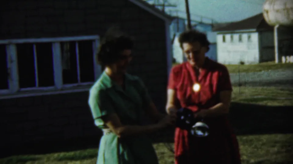 Two women in green and red dresses stand together on a Chadron lawn, with one holding a camera in 1959 8mm footage.