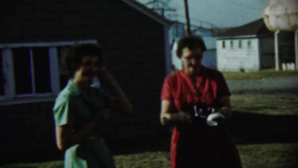 A woman in red holds a camera as a woman in green adjusts her hair on a Chadron, Nebraska lawn in 1959 footage.