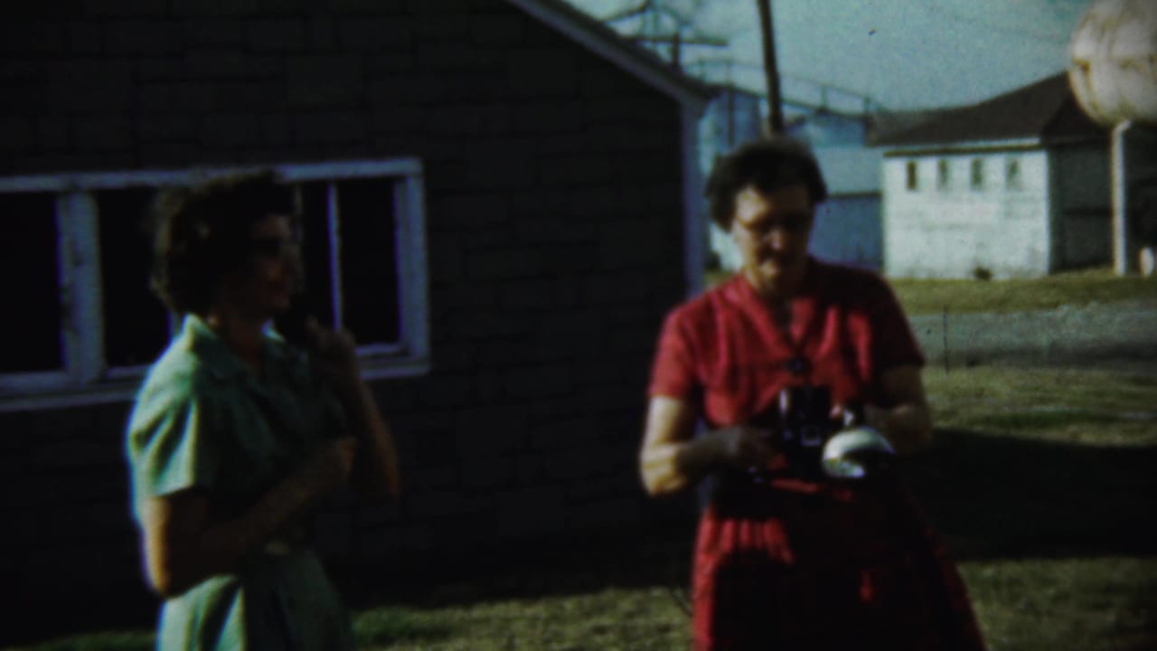 Two women stand outside a brick house, one holding a camera.