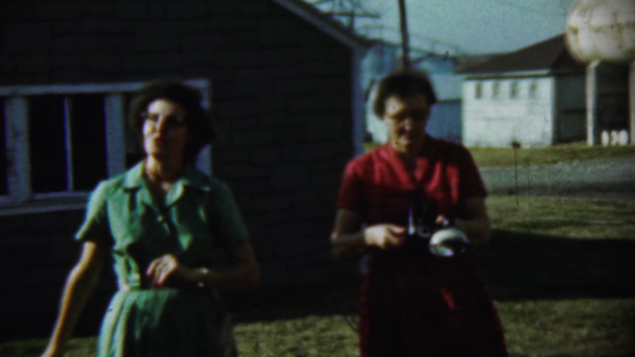 A woman in a green dress faces the filmer as another woman in red looks down at a camera outside a Chadron house in 1959.
