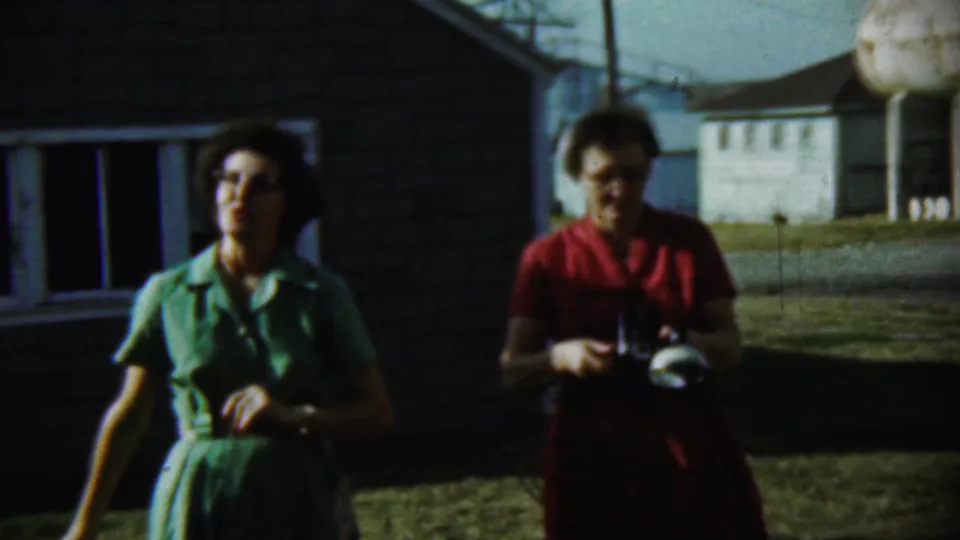 A woman in a green dress faces the filmer as another woman in red looks down at a camera outside a Chadron house in 1959.