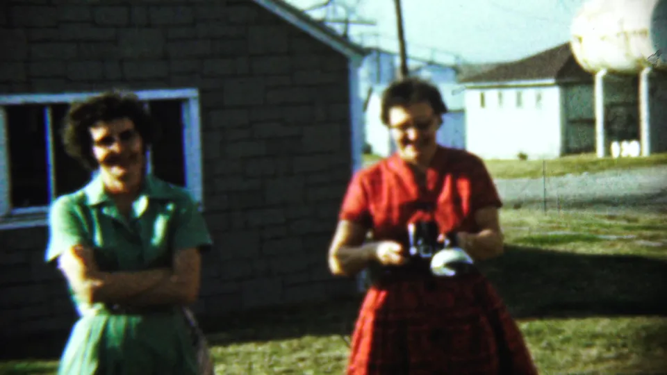 Two women pose near a brick house in Chadron, with the woman in green crossing her arms and the woman in red holding a camera.