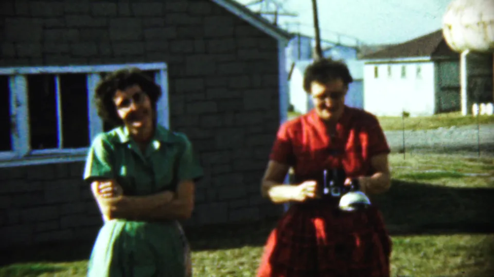 At the end of the clip, two women stand outside a Chadron brick house, one smiling with arms crossed and the other holding a camera.
