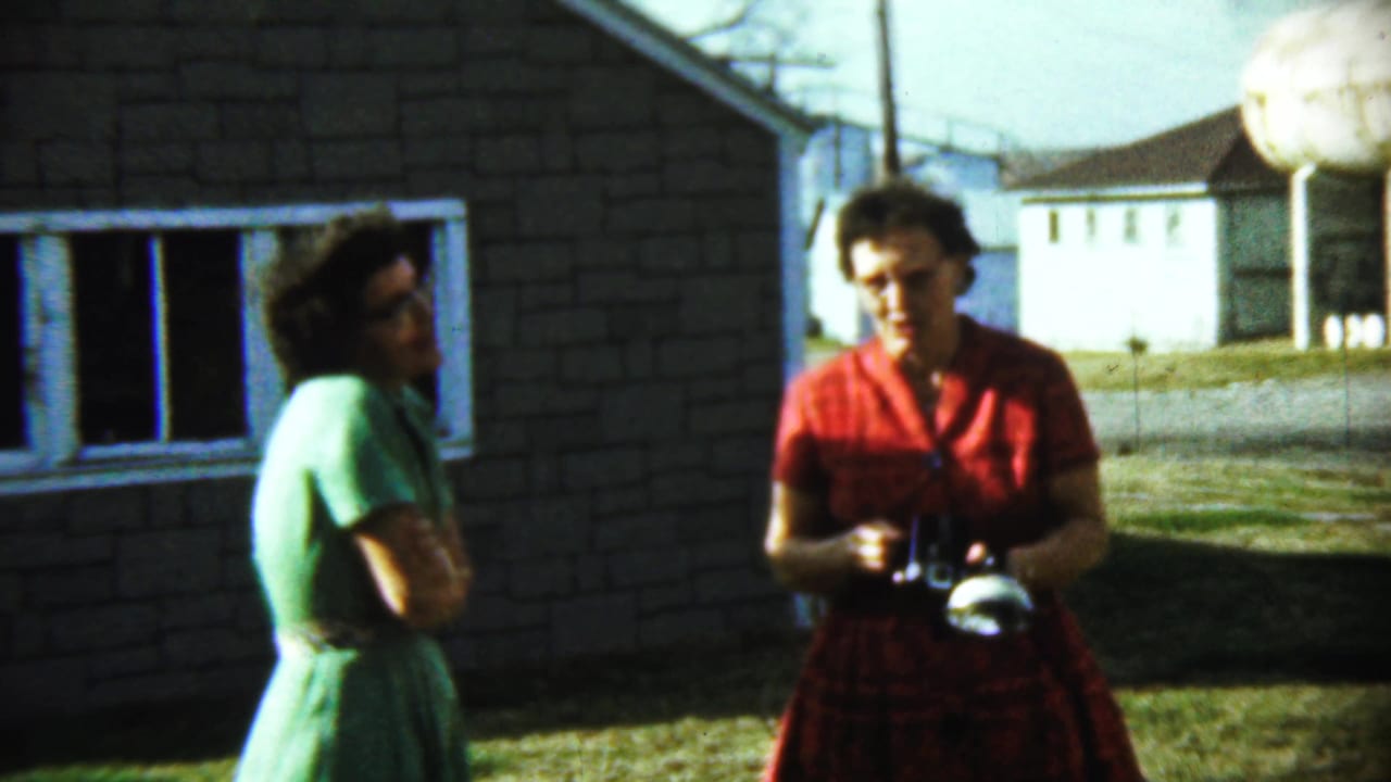 Two adult women stand outside a brick house, one in green and one in red holding a camera.