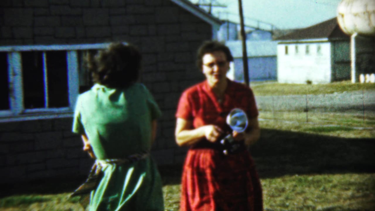 Woman in green faces away while woman in red stands with a camera outside the house.
