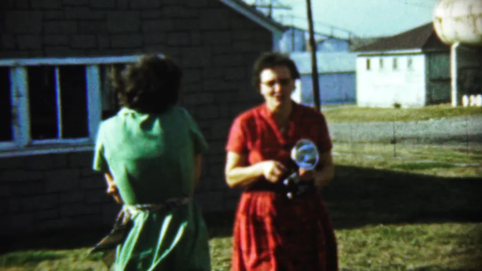 A 1959 Chadron home-movie frame shows the woman in green turned away and the woman in red holding a camera near a brick house.
