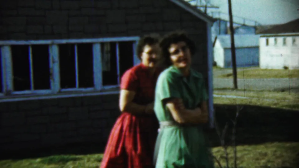 Two adult women in 1950s dresses stand close together outside a brick house in Chadron, Nebraska, looking toward the home-movie camera.