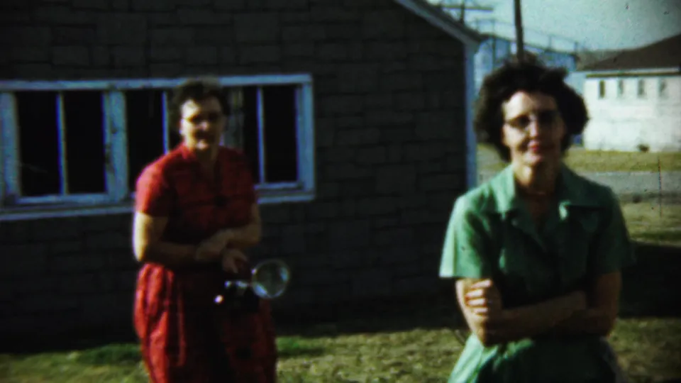 The woman in green stands nearer the camera with folded arms while the woman in red, holding a camera, remains by the brick house in Chadron.