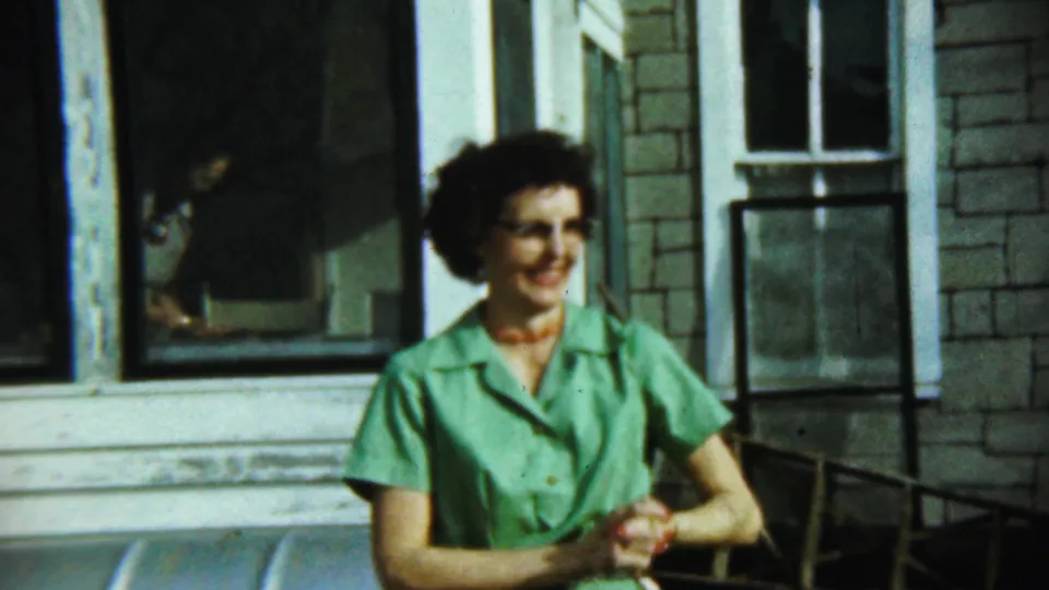 A woman in a green dress and glasses smiles outside a house in Chadron, Nebraska, with another figure partly visible nearby.