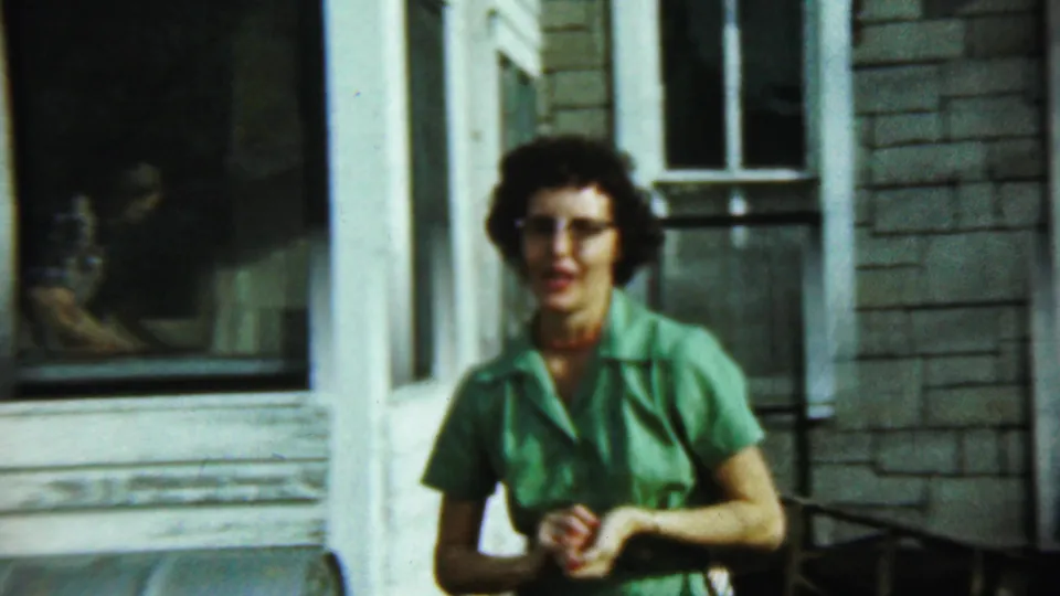The woman in green faces the camera near the house facade, with porch trim and windows filling much of the frame.