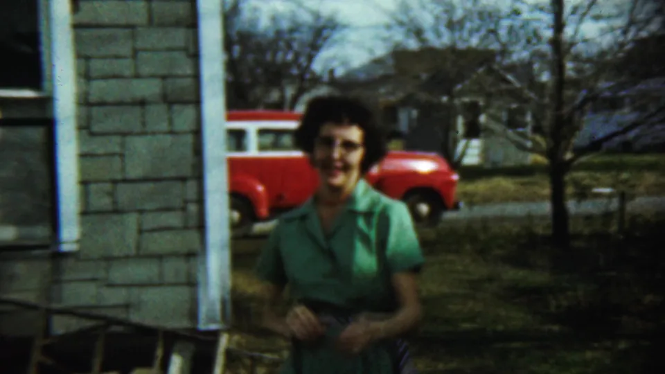 The woman in green stands in the yard with a red-and-white station wagon and bare trees visible in the background.