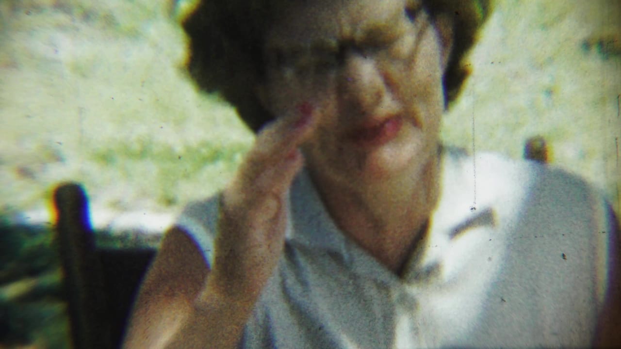 Elderly woman in glasses and a white sleeveless collared top raises her hand near her face outdoors.