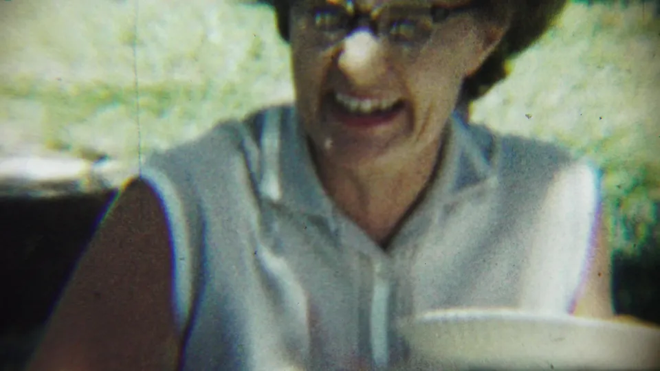A smiling elderly woman in glasses appears in soft 8mm focus at the Chadron, Nebraska picnic, with a plate visible in front of her.