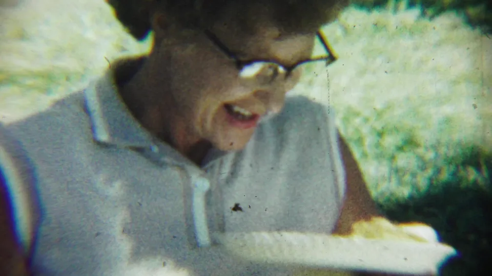 At the 1959 Chadron picnic, an elderly woman in glasses looks down toward a plate of food in bright, grainy 8mm footage.