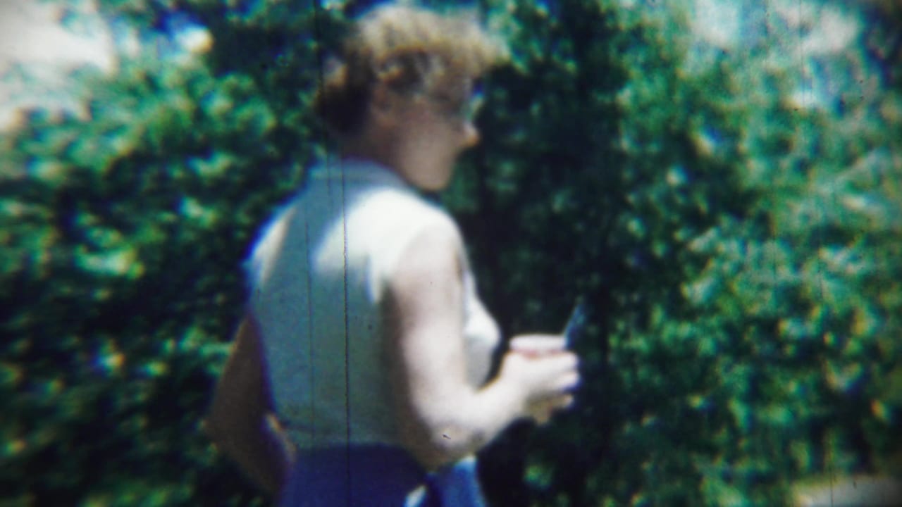 Woman seen from behind holding a small red cup or object near trees.