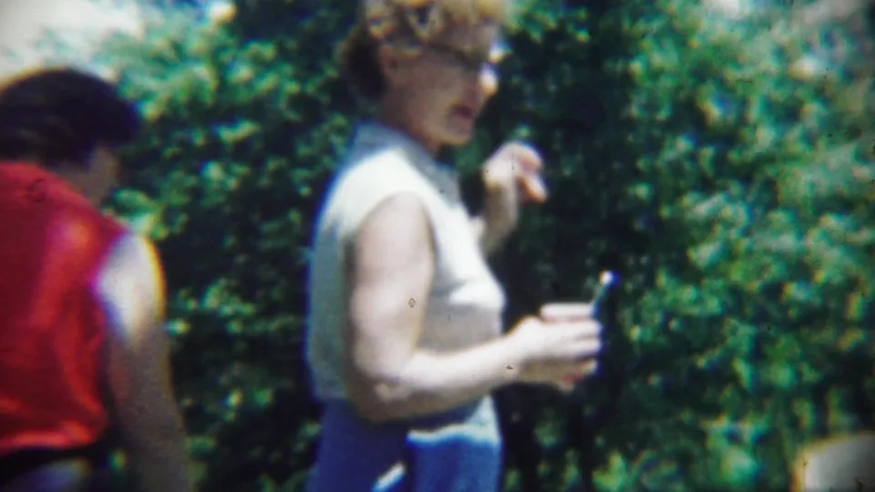 A woman in a white sleeveless top gestures while holding a small cup or object, with a red-clad person partly visible at the edge.