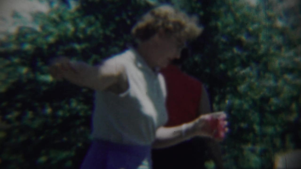 Woman in a sleeveless light top and blue skirt walks past greenery holding a red cup.