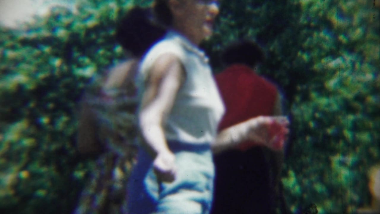 A woman in light summer clothing passes through a leafy Chadron garden area, with other adult attendees partly visible behind her.
