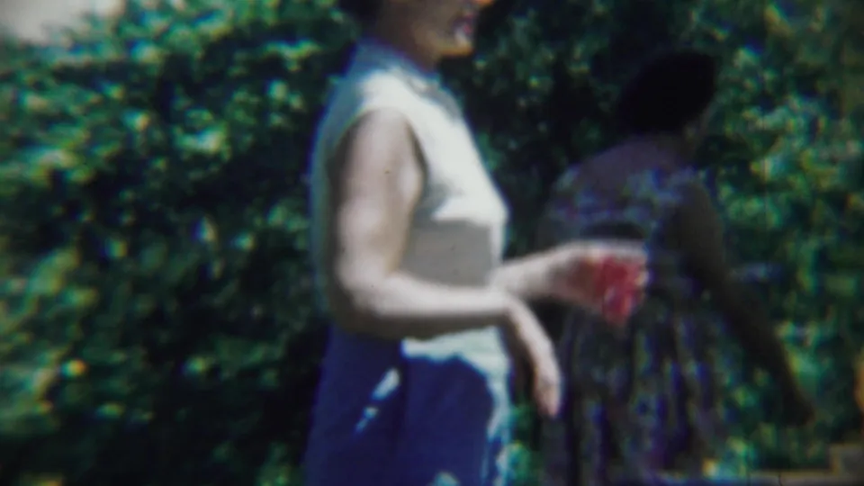 A woman holding a red cup crosses the foreground as another adult in patterned clothing moves through the Chadron garden setting.