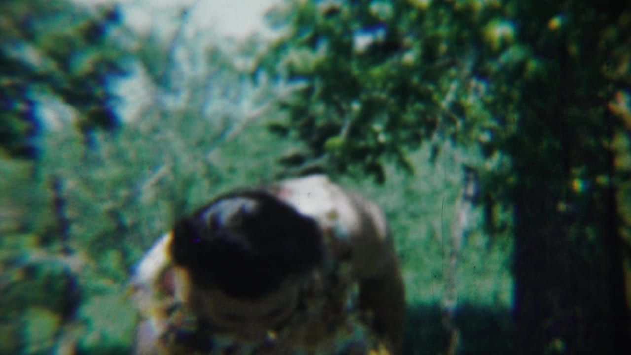 Woman in a floral dress bending near trees and green foliage.