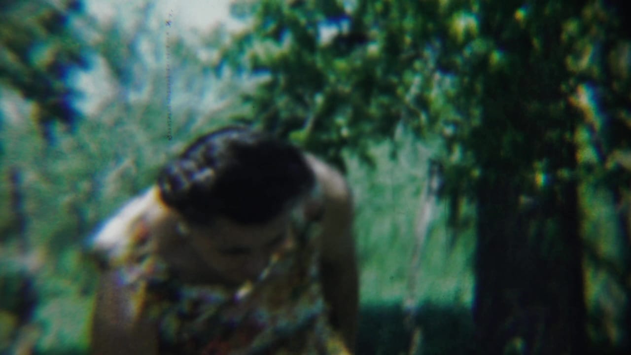 The woman leans forward near a tree, surrounded by tall grass and leafy branches in the 1959 Chadron home-movie scene.