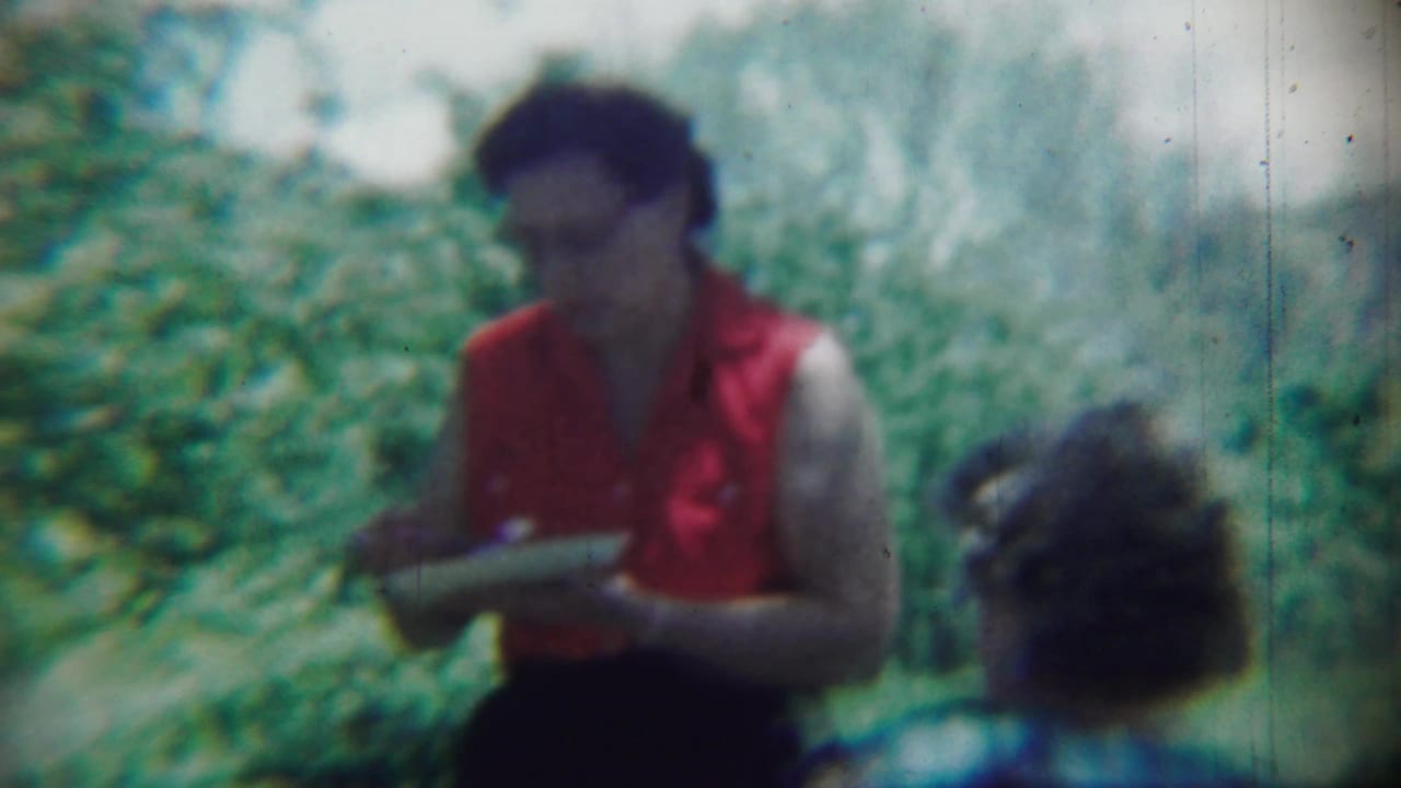 Woman in a red sleeveless blouse holding a plate outdoors near green foliage.