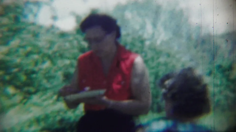 In 1959 Chadron home-movie footage, a woman in a red sleeveless blouse stands with a plate amid dense greenery.