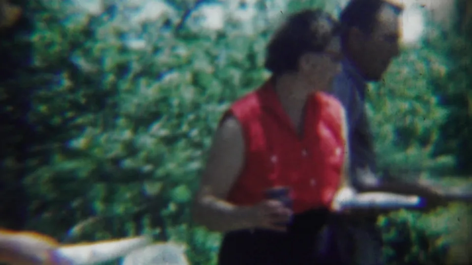 At the outdoor gathering, the woman in red holds a cup and plate as a man in a blue shirt stands partly behind her.