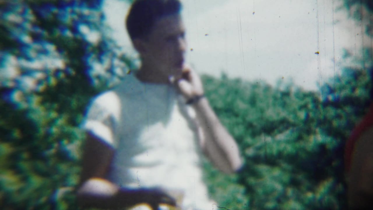 A man in a white T-shirt appears to eat or drink outdoors while a person in red stands beside him in the 1959 Chadron footage.