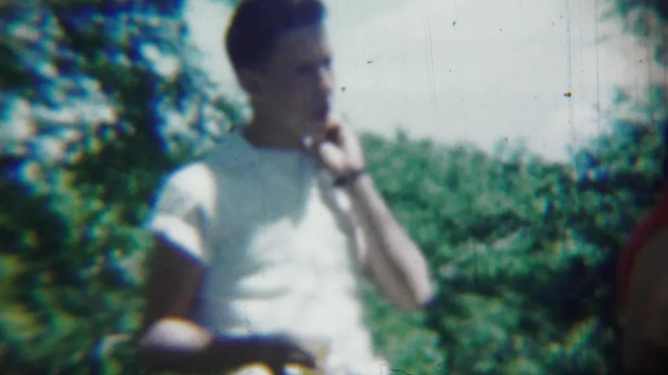 A man in a white T-shirt appears to eat or drink outdoors while a person in red stands beside him in the 1959 Chadron footage.