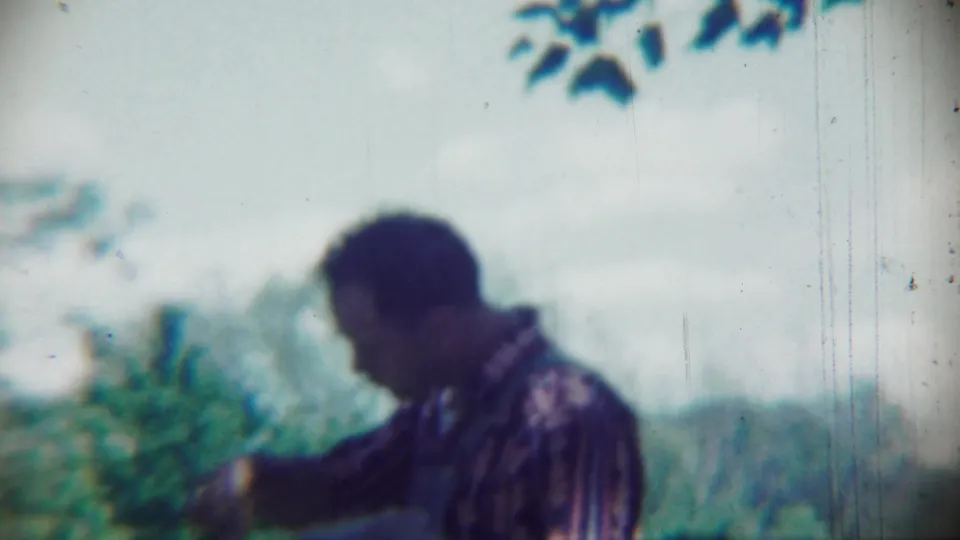 An adult man in a patterned shirt looks down in soft-focus 8mm footage from Chadron, Nebraska, with trees behind him.