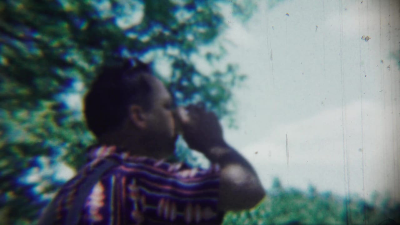 Man in a striped shirt drinks from a cup outdoors beneath leafy trees.