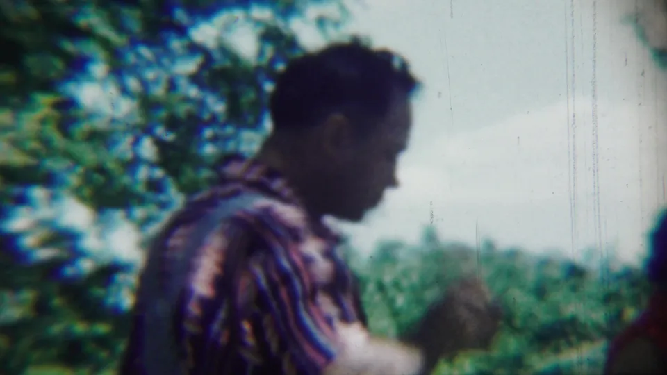 A man in a striped shirt lowers his hand after drinking, with leafy trees and distant greenery visible in the home-movie frame.