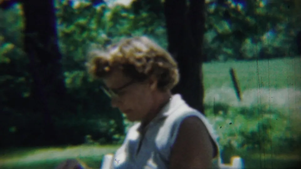 An adult woman with glasses appears in a sunny garden-like setting, with trees, lawn, and greenery behind her in the 1959 home movie.
