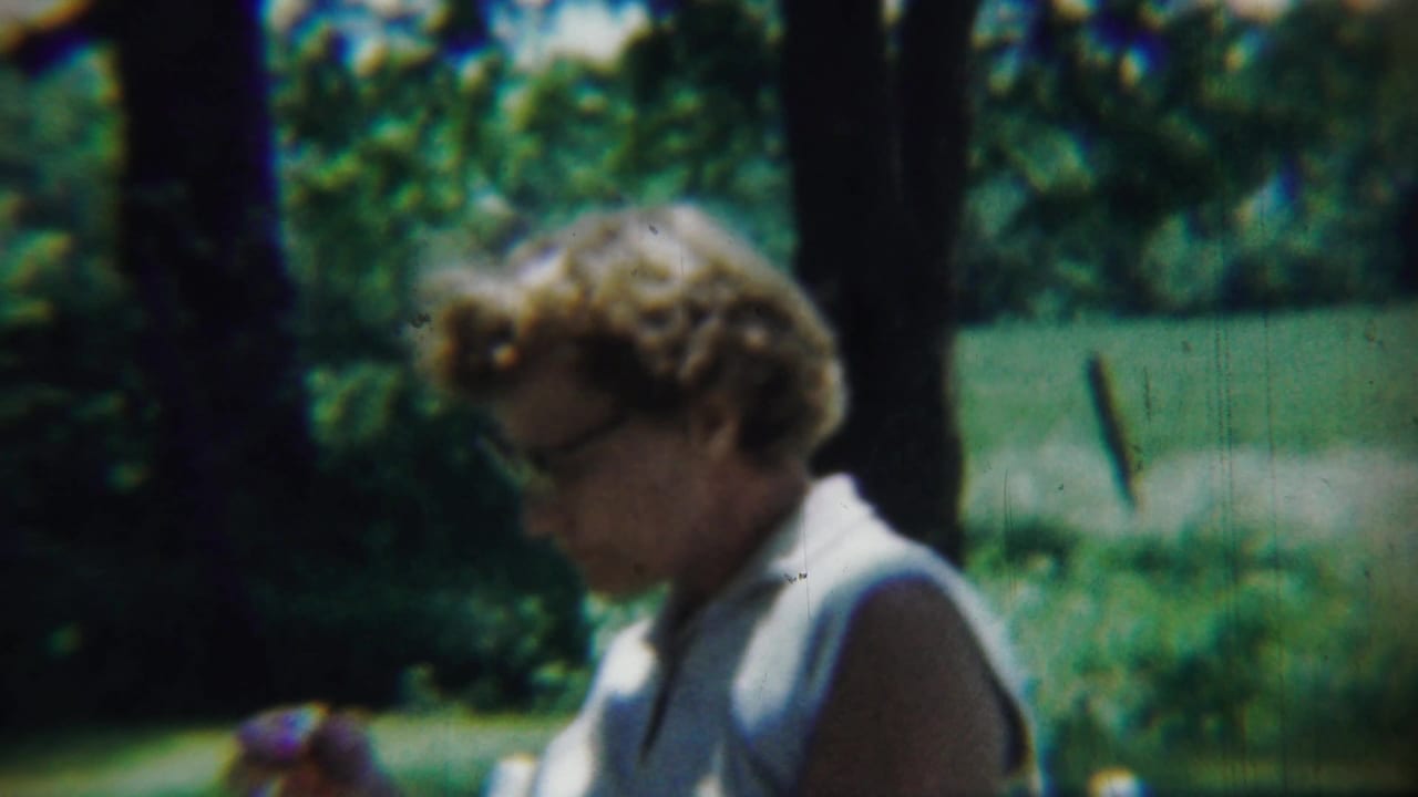 In 1959 Chadron, Nebraska footage, a woman with glasses sits outdoors in a white sleeveless top amid trees and grass.