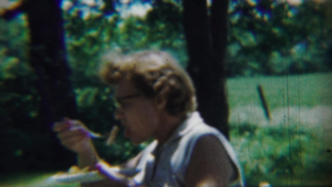 Woman in glasses raises a fork while seated outdoors with trees and grass behind her.