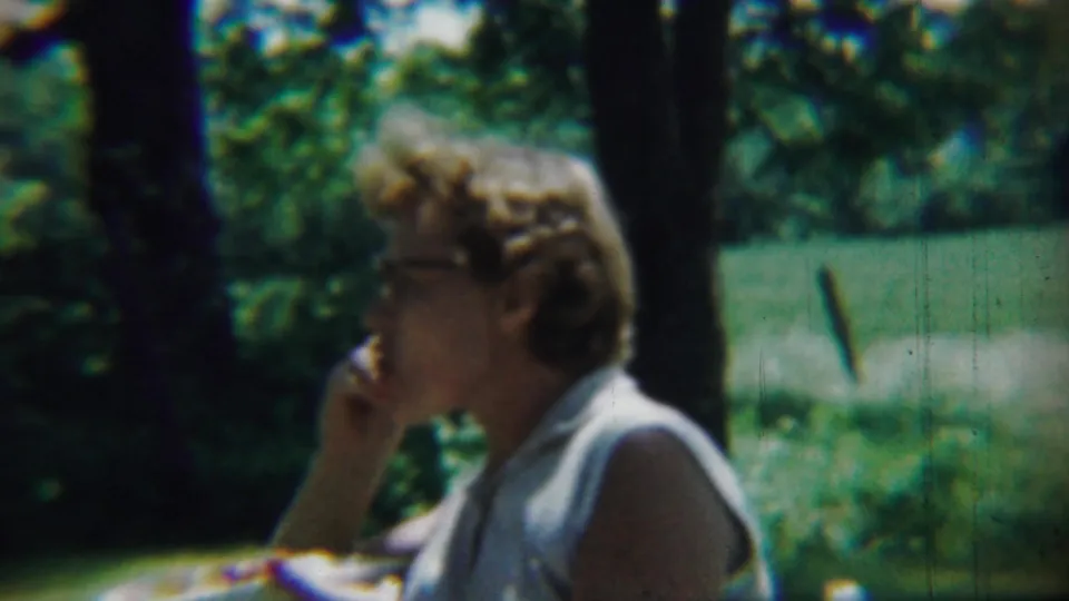 The 1959 Chadron, Nebraska home-movie frame shows a woman with glasses eating in a shaded, leafy outdoor setting.