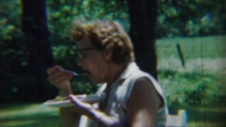 In a soft 8mm frame from 1959 Chadron, Nebraska, a woman with glasses eats from a plate in a green outdoor area.